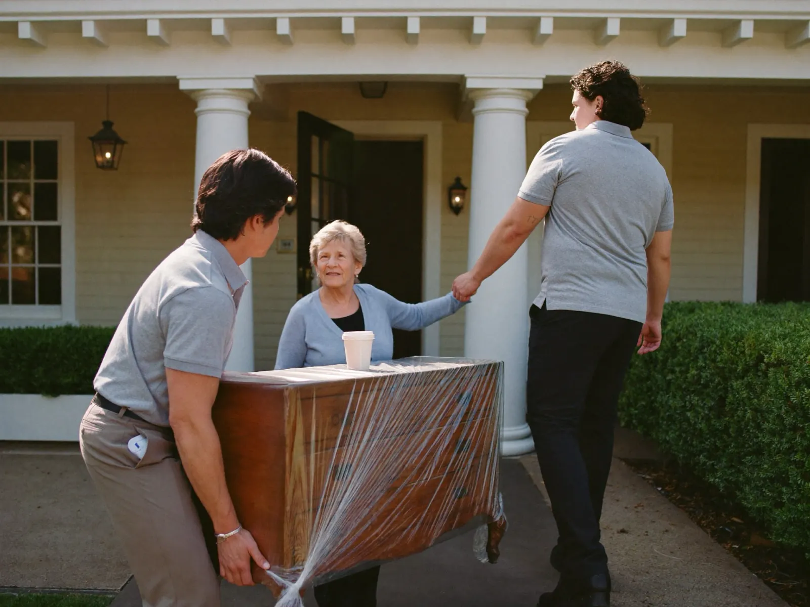 Two movers carrying a wrapped dresser toward a homeowner waiting on her porch